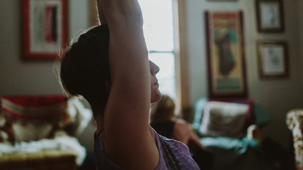 Female practising yoga, viewed at close range from the side, arms raised above her head, a lounge setting is in the background